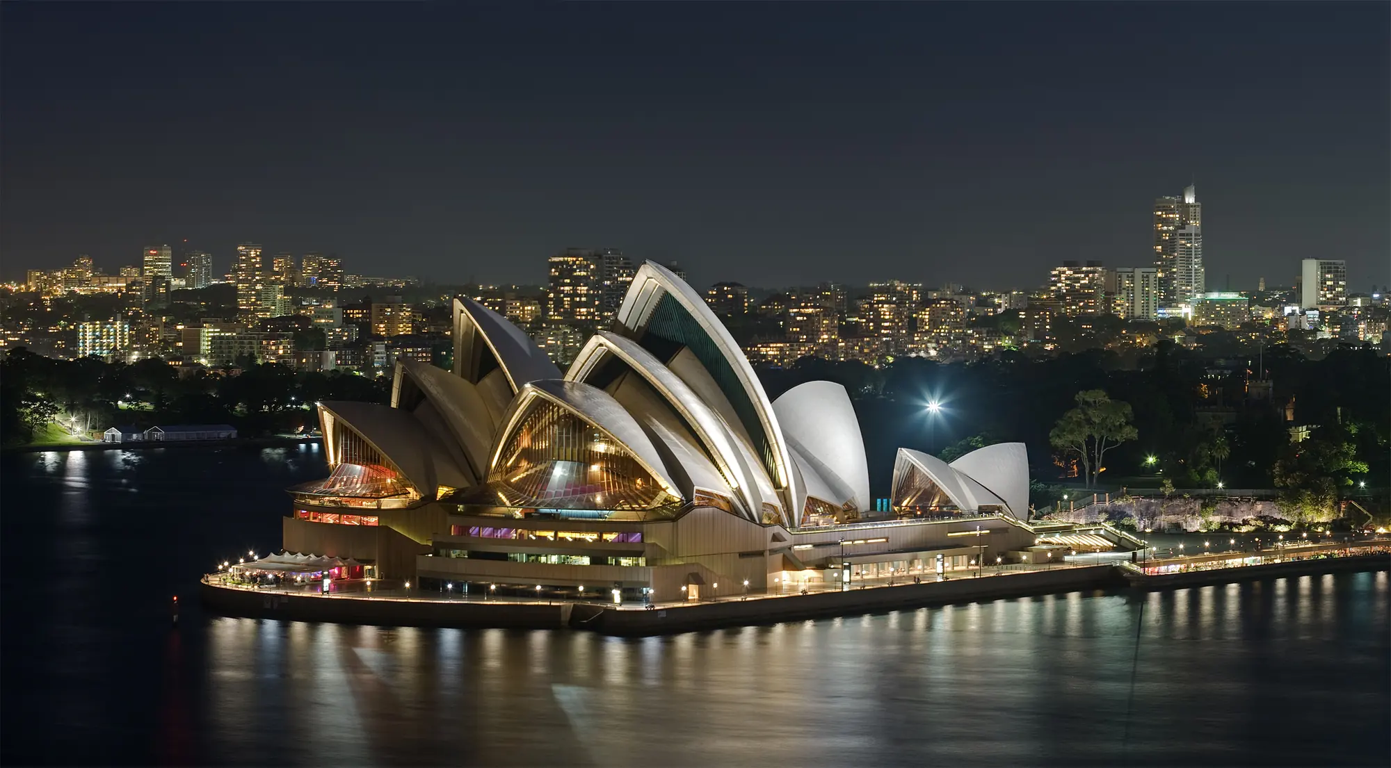 The Sydney Opera House lit at night on Bennelong Point, seen across the water, with the city lights behind