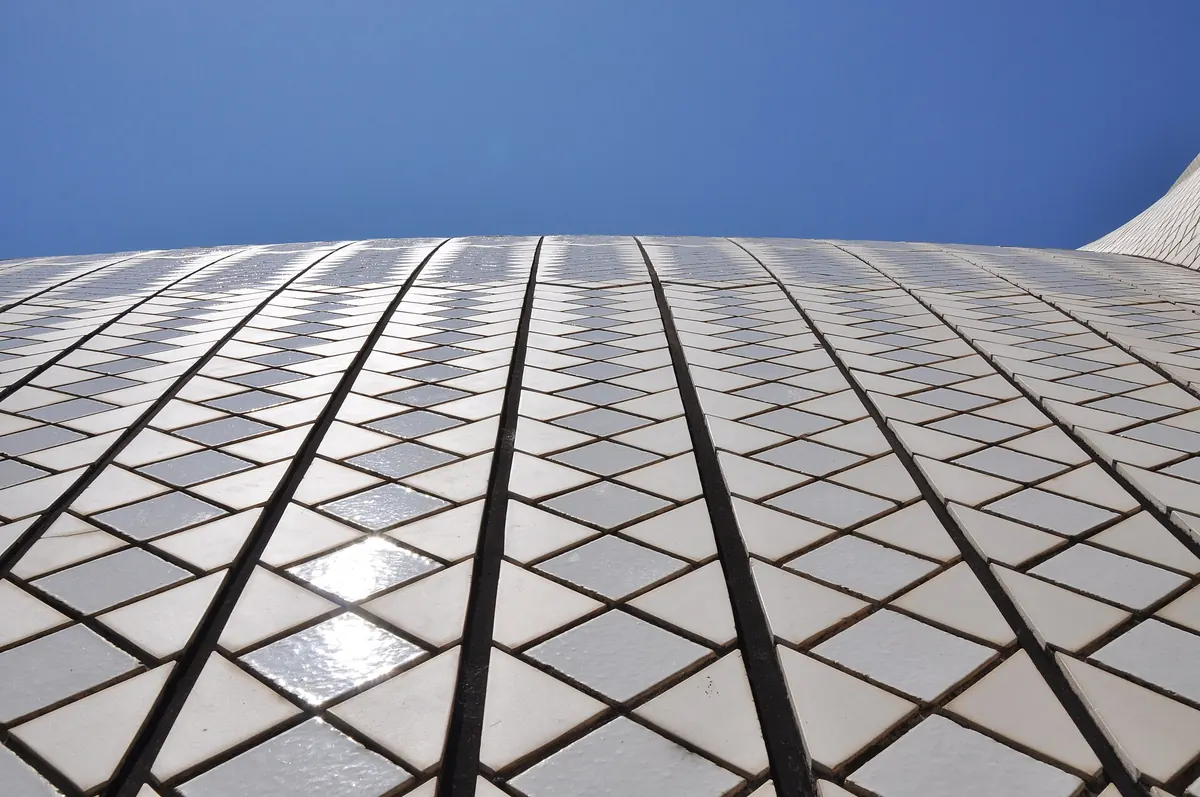 Looking up the white tiled surface of an Opera House shell against a clear blue sky, the tiles laid in chevrons