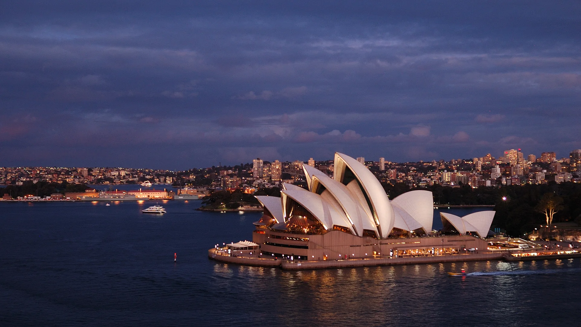 The Sydney Opera House at dusk, its white sails lit above the dark water of Sydney Harbour, with the city lights behind